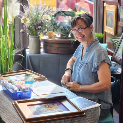Ellen Paquette is sitting at a table with art supplies and framed artwork in a studio setting.