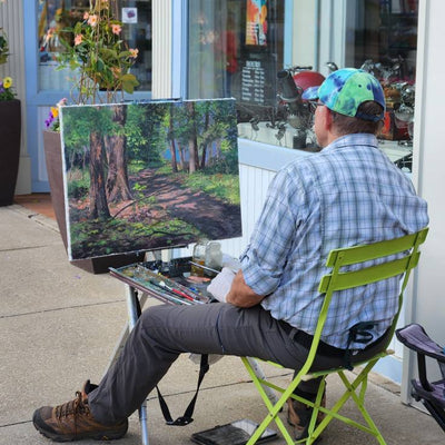 Jack Paluh is painting an outdoor scene on an easel in front of the Painted Finch Gallery store window.